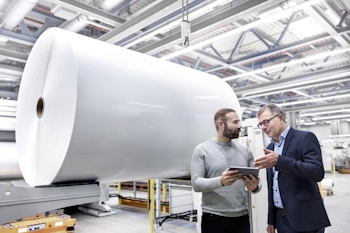 Two men in a factory are discussing a tablet while a large roll of paper stands in the background.