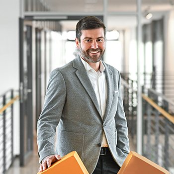 A man in a gray blazer smiles and holds two orange objects in a modern office setting.