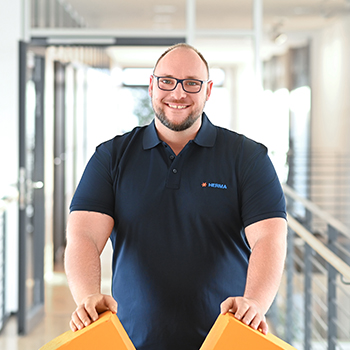 A man wearing glasses and a polo shirt stands in a modern office, holding two orange objects in his hands.