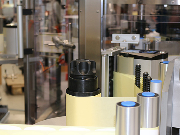 A black bottle sits on a conveyor belt in a production facility, surrounded by machines and equipment.