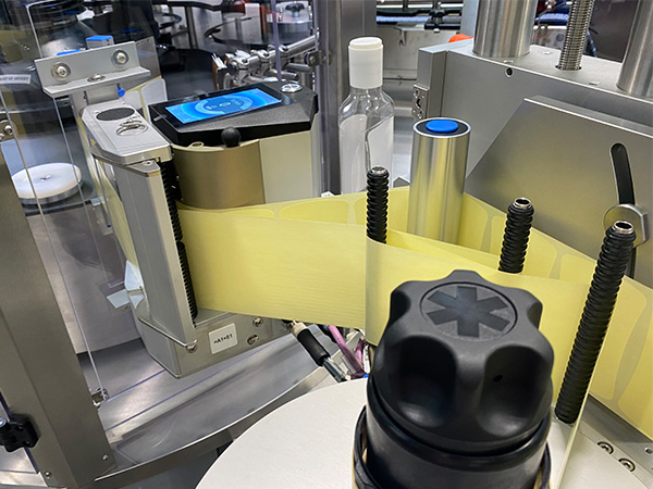A labeling device with a yellow label, which is applied to bottles, is located in a production environment.