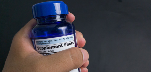 A hand is holding a blue bottle of dietary supplements, with the supplement facts visible on the label.