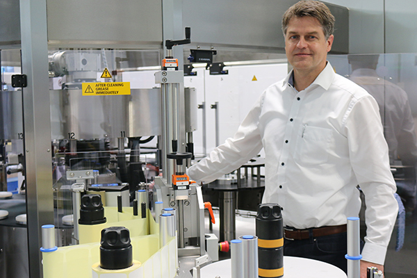 A man stands next to an industrial machine for processing bottles, surrounded by various containers and technical equipment.