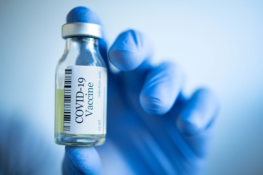 A hand in blue gloves holds a glass bottle labeled 'COVID-19 Vaccine.