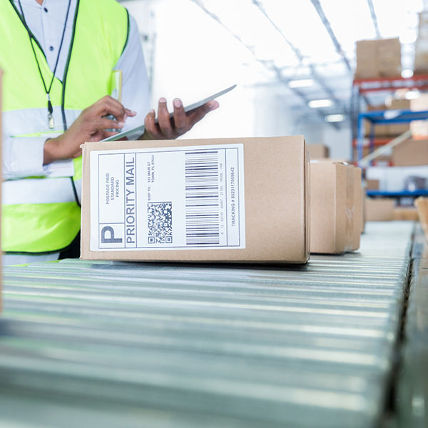 A staff member in a safety vest is checking packages on a conveyor belt while holding a smartphone in hand.