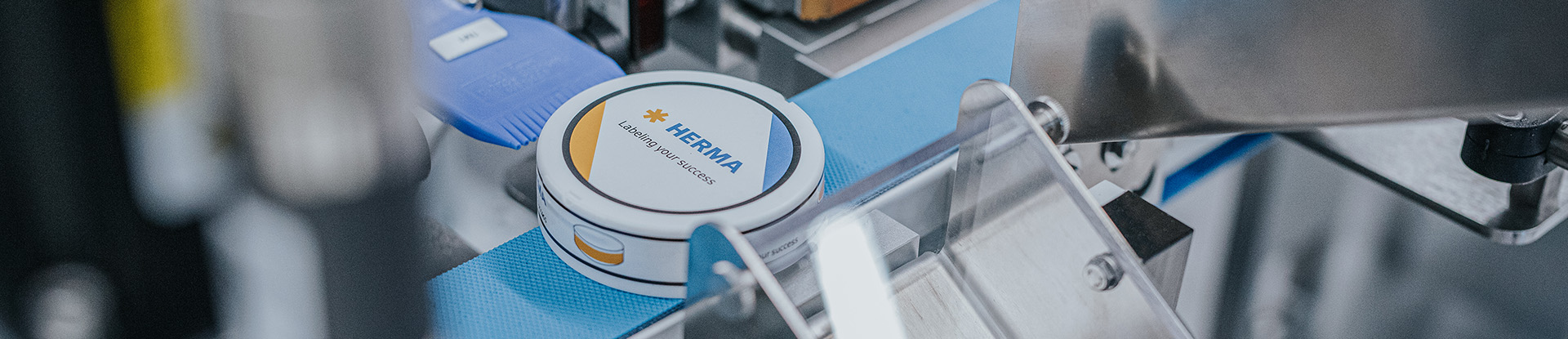 A can with the 'HERMIA' logo on a conveyor belt in a production environment.