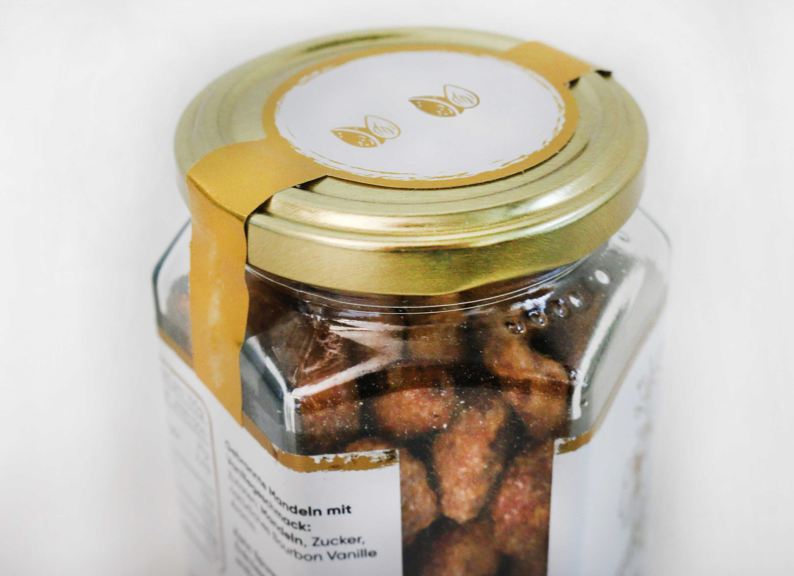 A glass jar with a gold-colored lid, filled with roasted almonds. On the lid, there is a label featuring a minimalist design.