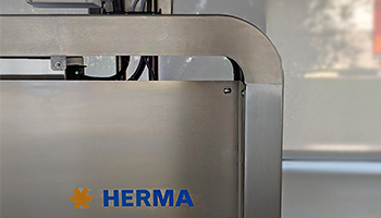 A part of a machine with the logo 'HERMA' in blue lettering on a metallic surface.