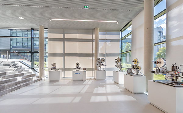 Modern machines and devices are displayed on white pedestals in a bright, airy room with large windows.