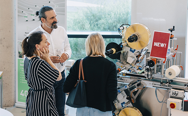 Three people are standing in front of a machine that applies labels. One person is explaining the functions of the machine while the others listen.