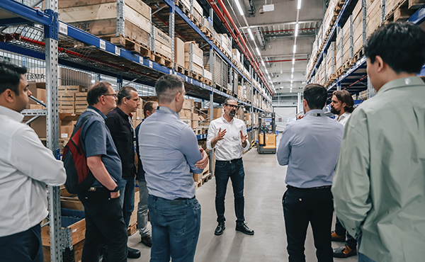 A group of people is listening to a speaker in a warehouse filled with shelves of boxes.