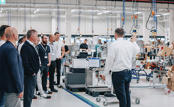 A group of people is listening to a speaker in a modern production hall, surrounded by machines and workstations.