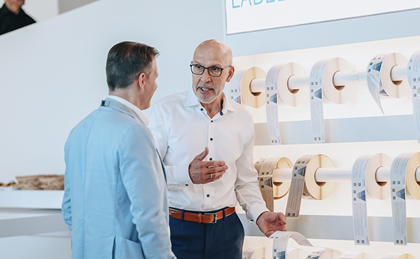 Two men are conversing in front of a wall covered with rolls of labels. The man in white is explaining something while the other listens attentively.