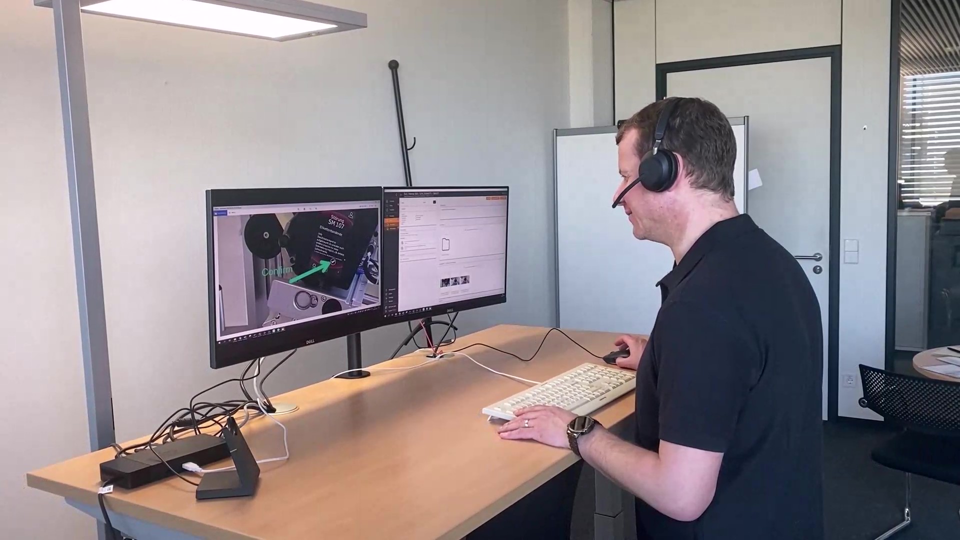 A man is sitting at a desk with two monitors, wearing a headset and working on a computer.