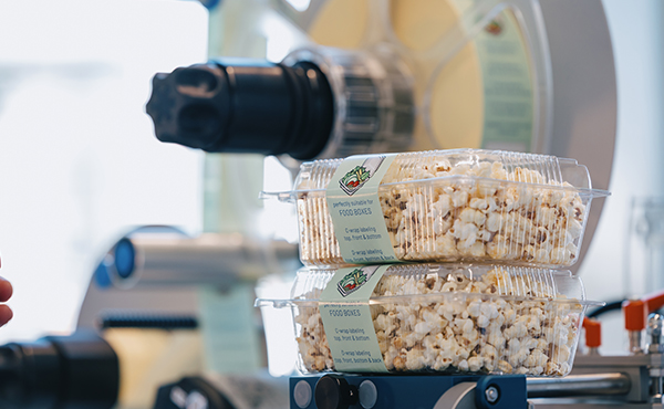 Two transparent containers filled with popcorn are stacked on top of each other, while a popcorn machine is visible in the background.