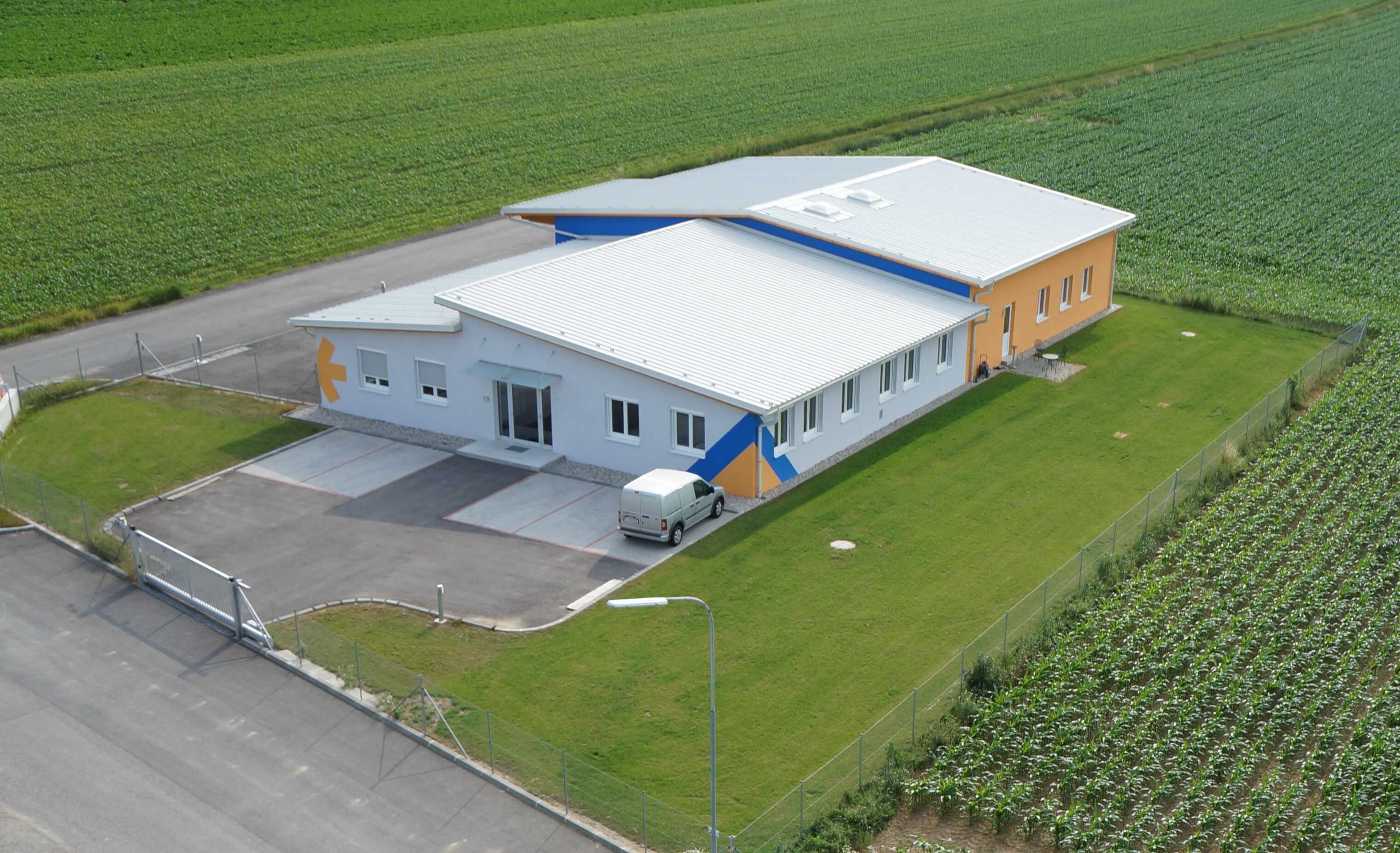 A modern building with a flat roof, a bright facade, and colorful accents, surrounded by agricultural fields.