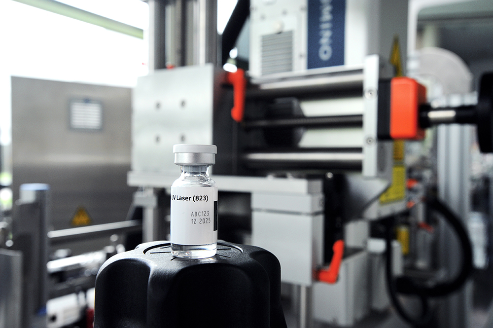 A syringe filled with a clear liquid is placed on a black lid in front of an industrial machine.