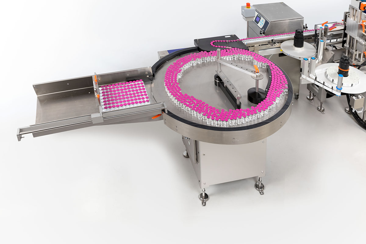 Automated filling system with a circular conveyor belt, filled with small bottles. Pink labels are visible.