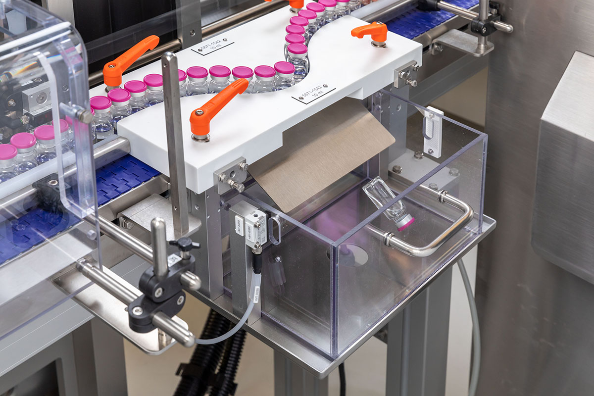 Automated filling system with bottles on a conveyor belt and a container for receiving packaging material.