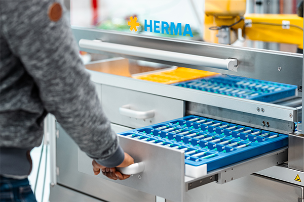 A person pulls out a drawer with blue compartments from a shelf marked with the logo 'HERMA'.