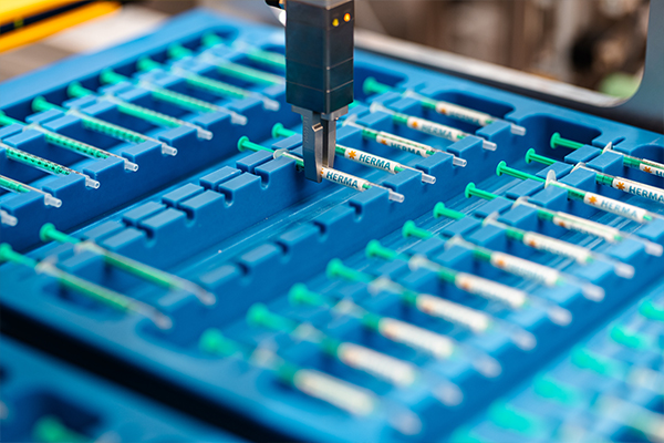 An automated device retrieves syringes from a blue container with several compartments designed for syringe storage.