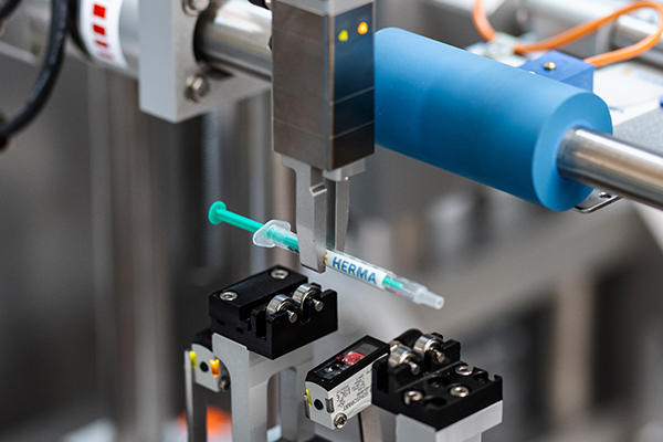 An automated gripper holds a syringe labeled 'HERMA' in an industrial setting.