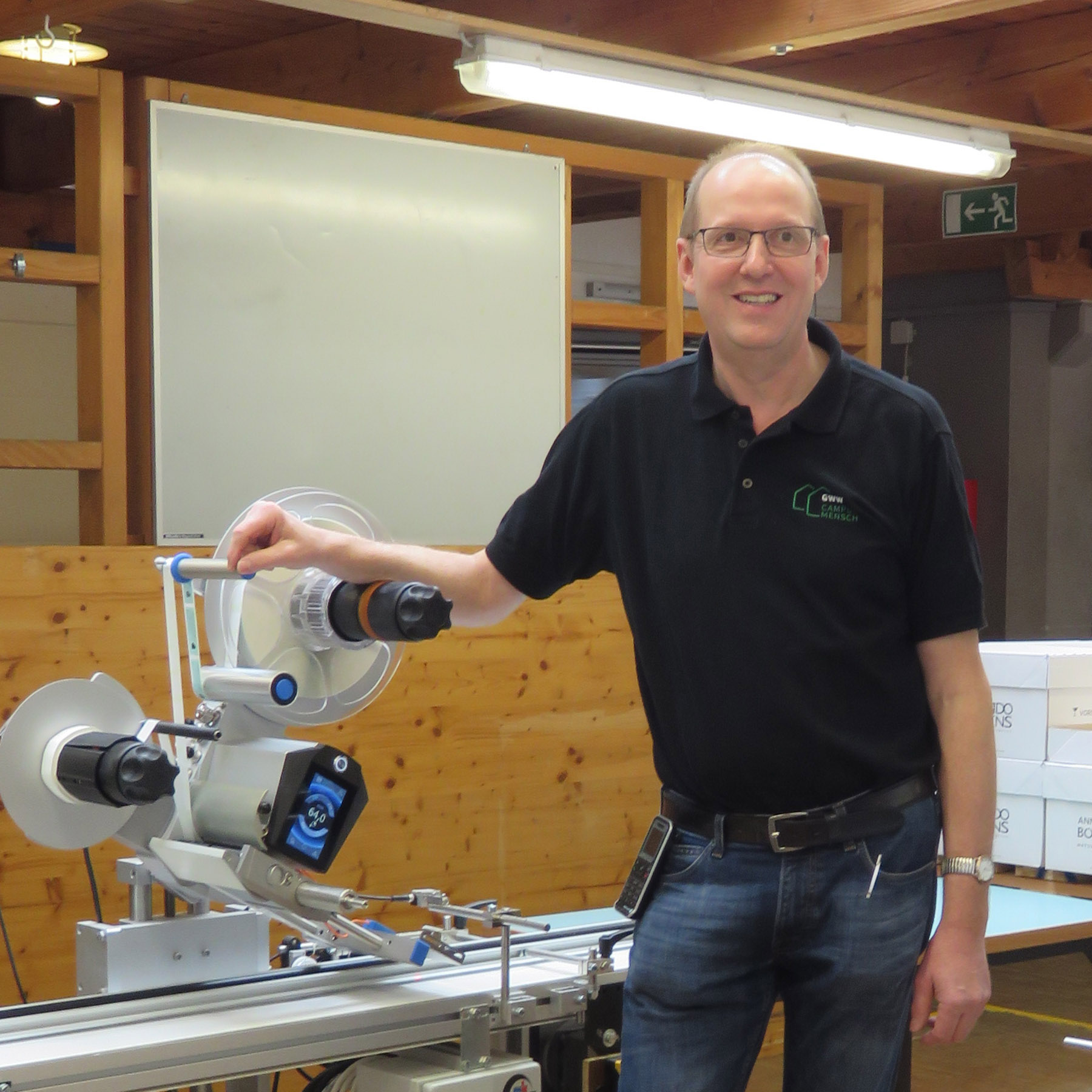 A man stands next to a machine in a workshop, smiling and wearing a black T-shirt with a logo.