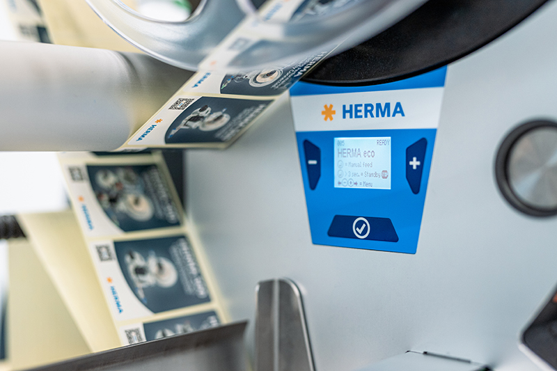 A label printer with a display showing the HERMA brand. Labels are dispensed on a roll.