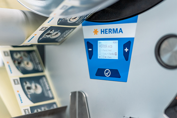 A label printer with a digital control panel displaying the brand 'HERMA.' The label rolls are visible.