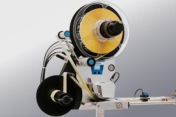 A machine component with a large spool of yellow material, connected with hoses and a control panel, for processing or packaging.