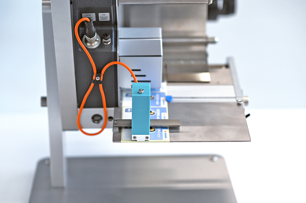 A technical device with a blue component mounted on a metal plate, connected by an orange cable.