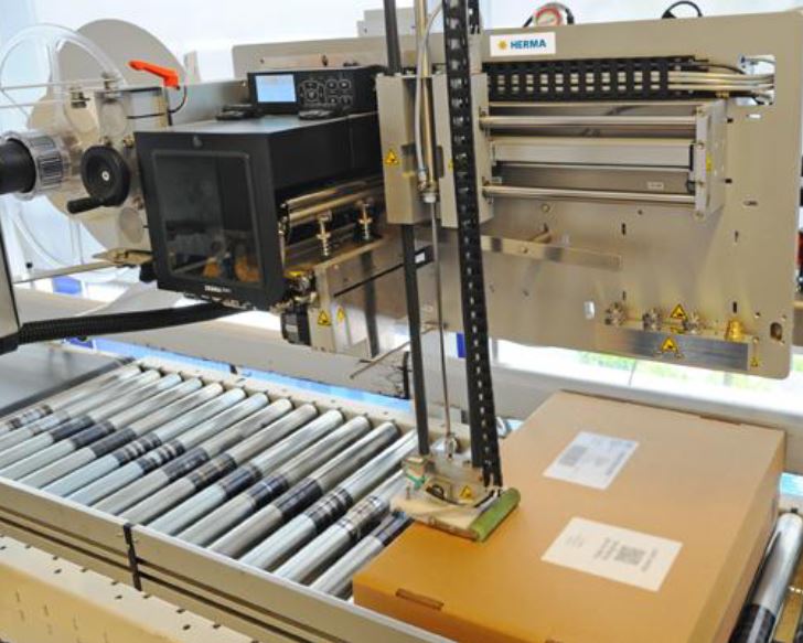 Automated labeling system with a box on a conveyor belt, equipped with printing and labeling mechanisms.