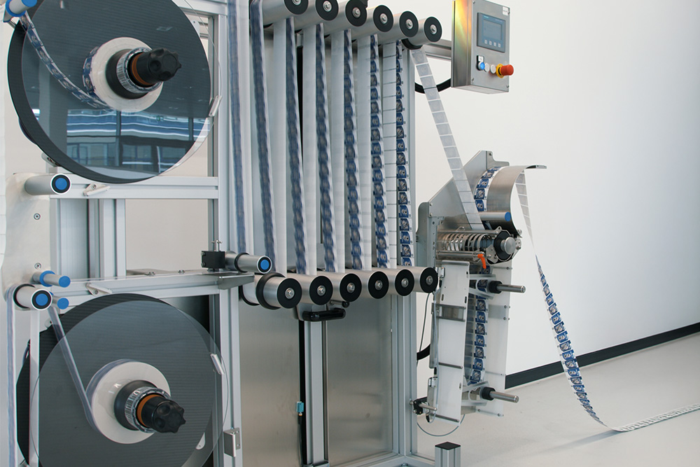 An industrial machine with two large spools that feeds a ribbon through several rollers, equipped with a control panel.