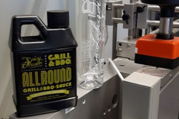 A bottle of barbecue sauce and a clear glass are placed on a machine in a production environment.
