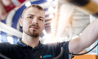 A man in a HERMA polo shirt is working on electrical cables in a modern setting with bright lighting.