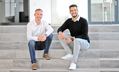 Two men are sitting on a staircase in a modern building. One is wearing a white shirt, while the other is in a black sweater.