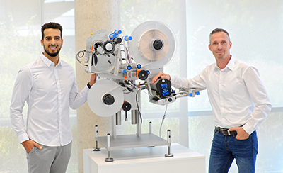 Two men in white shirts stand next to a technical machine with rollers and a display, designed for industrial applications.