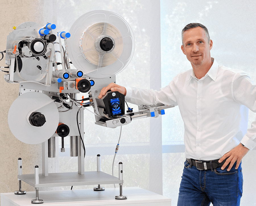 A man stands next to a modern labeling machine, holding a control device in his hand. The machine has several rollers and control elements.
