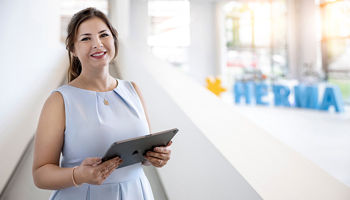 A woman in a light blue dress is holding a tablet and smiling. In the background, large, colorful letters spell out the word 'HERBIA.