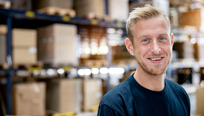 A smiling man with blonde hair stands in a warehouse with shelves full of boxes in the background.