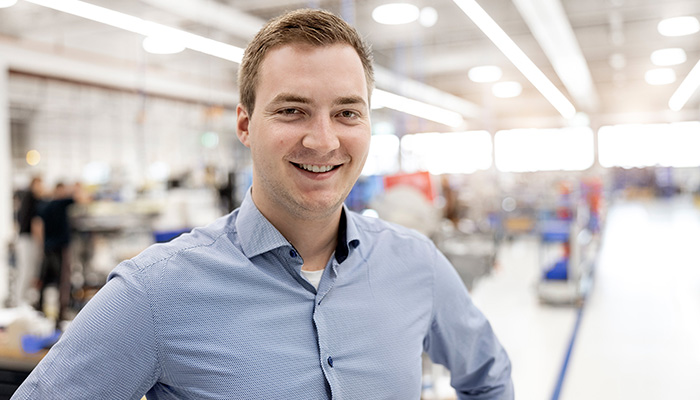 A young man in a light blue shirt smiles as he stands in a modern factory hall, surrounded by machines and workstations.