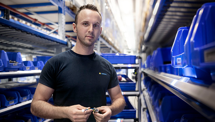 A man in a black T-shirt is holding a tool, surrounded by blue storage boxes on a shelving unit.