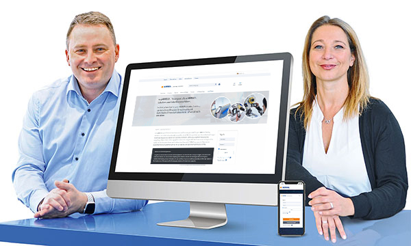 Two people are sitting at a table, next to a computer and a smartphone, both displaying a website.