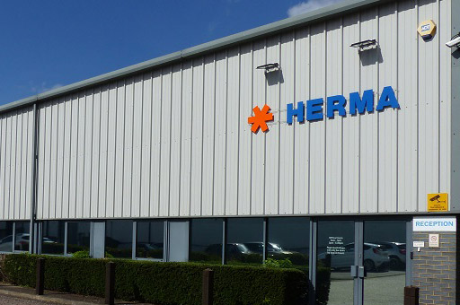 Facade of a modern building featuring the inscription 'HERMA' in blue lettering and an orange symbol. Reception area visible.