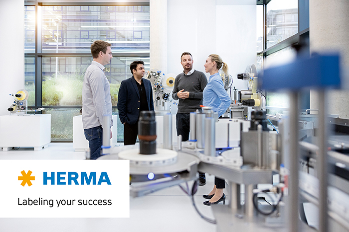 Four people are discussing in a modern room, surrounded by machines and equipment for labeling. A HERMA logo is visible.