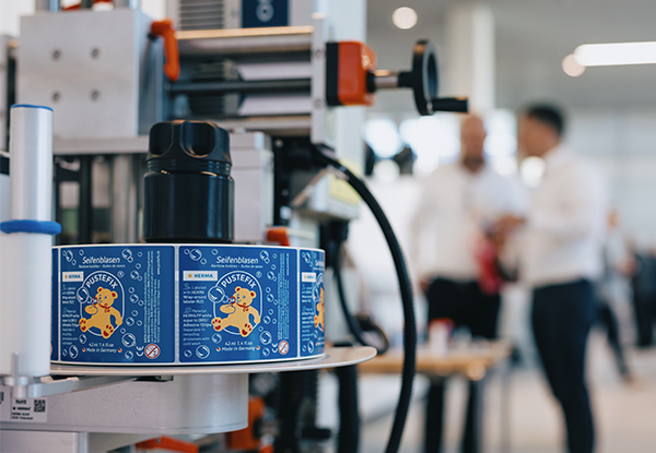 A packaging machine with colorful labels on cans in the foreground, while people are talking to each other in the background.