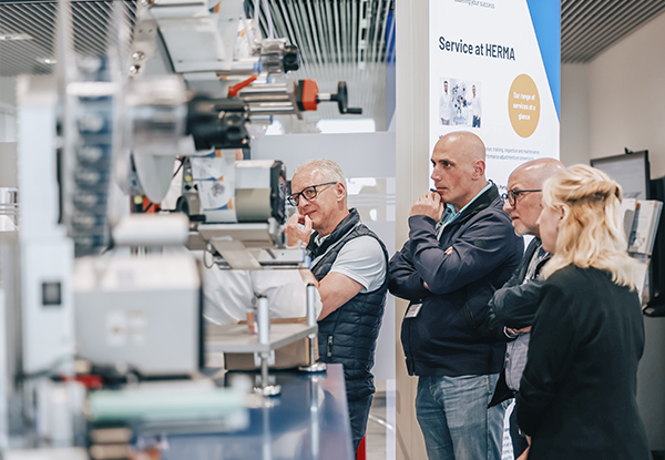 Four people are attentively watching a machine presentation at a trade fair, while information about the service at HERMA is being displayed.