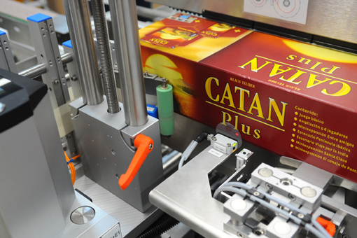 A packaging machine that processes a box of the game 'Catan Plus,' with various mechanical components in the foreground.