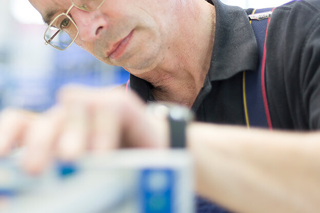 A man is working intently on a technical device in a workshop with blue storage boxes in the background.