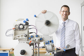 A man in a shirt and tie stands next to a technical machine with rollers and control elements.